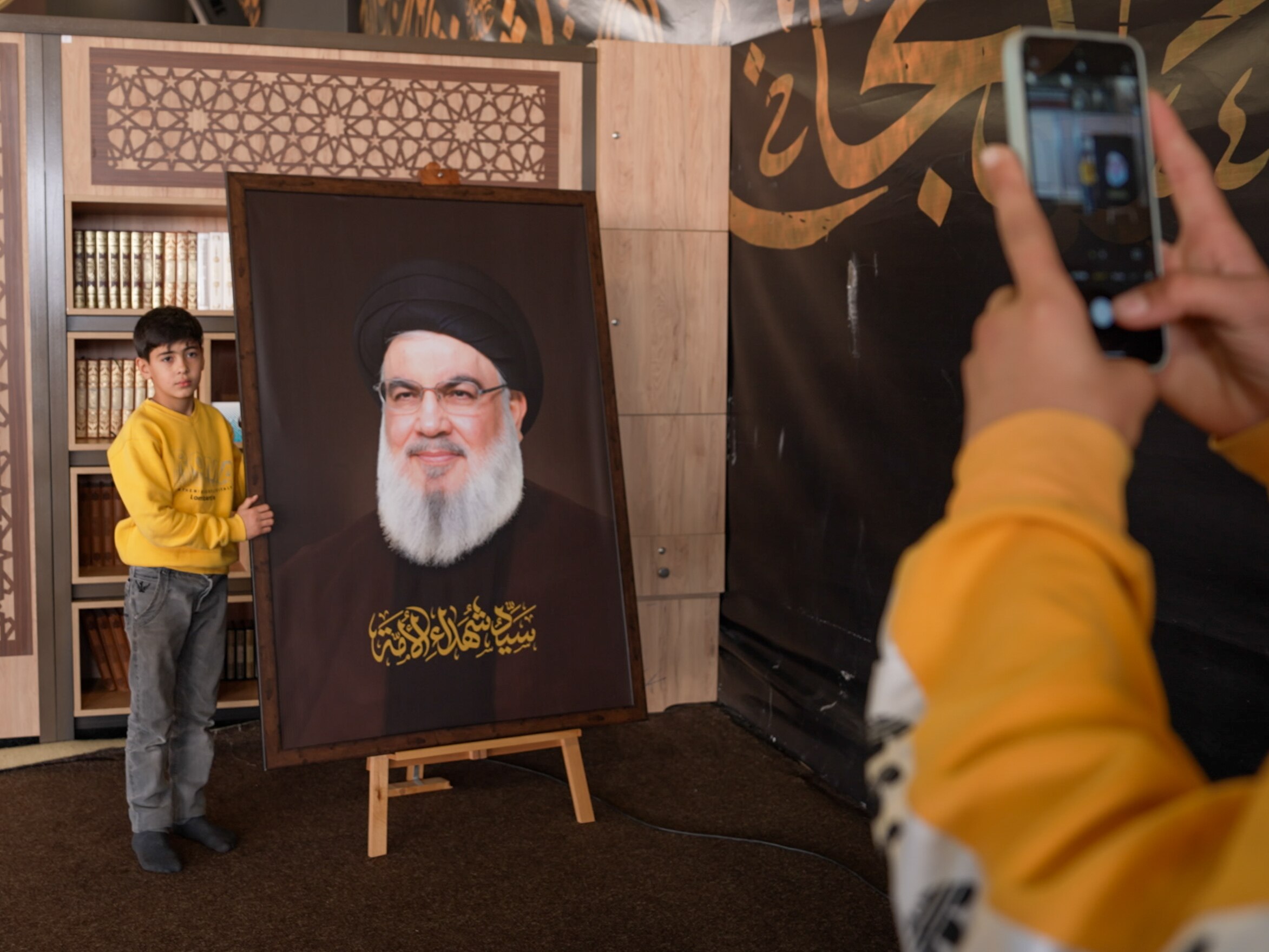 A child standing next to a large portrait of Hassan Nasrallah, getting his photo taken.