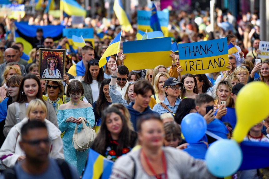 Толпа людей, в основном молодых женщин, идет вместе в сине-желтой одежде и с раскрашенными лицами.