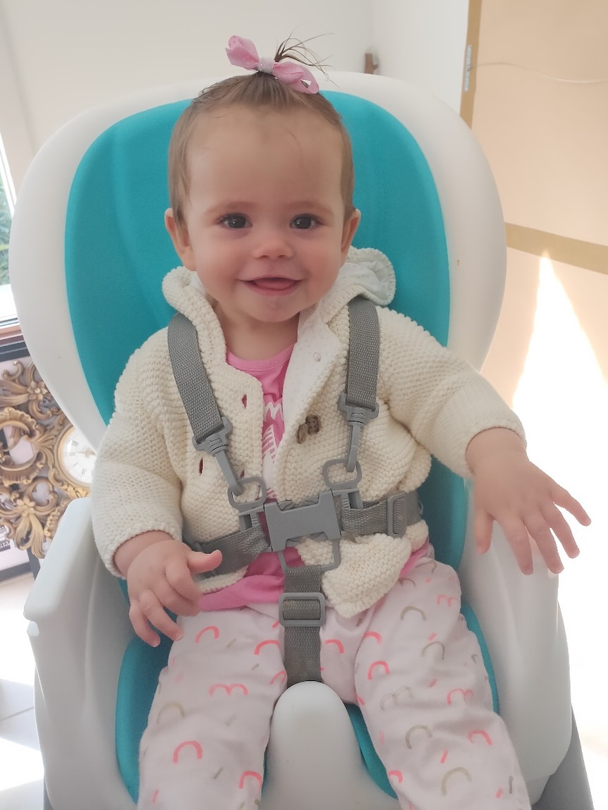 A baby wearing a pink hair tie sits in a blue and white high seat smiling at the camera