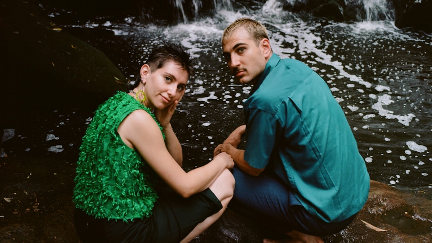 Niki and Solly crouch on wet stone in front of a waterfall, looking to the camera