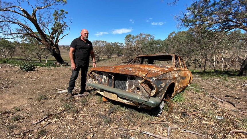 A man standing next to a burnt-out old car among scrubby trees