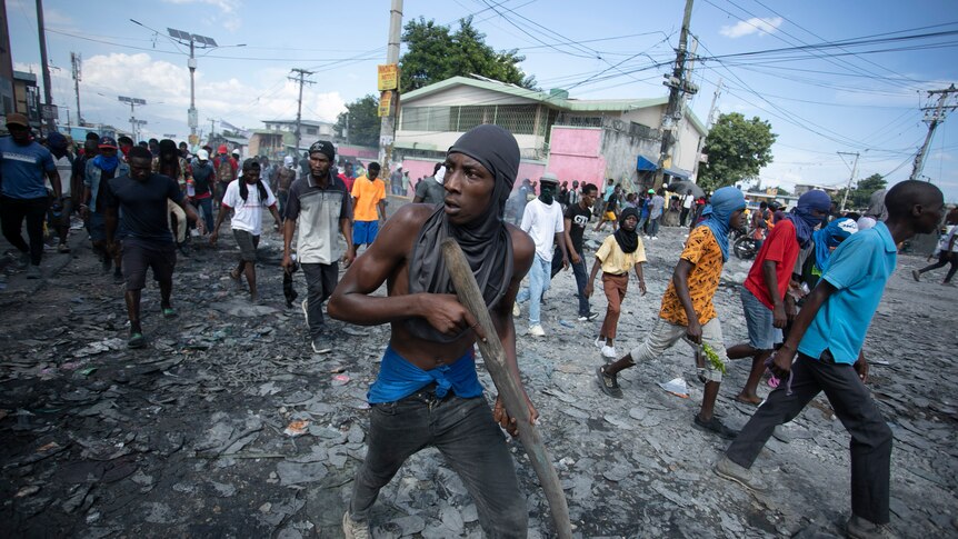a protester with a piece of material wrapped around his head carries a piece of wood as he walks with a group of men