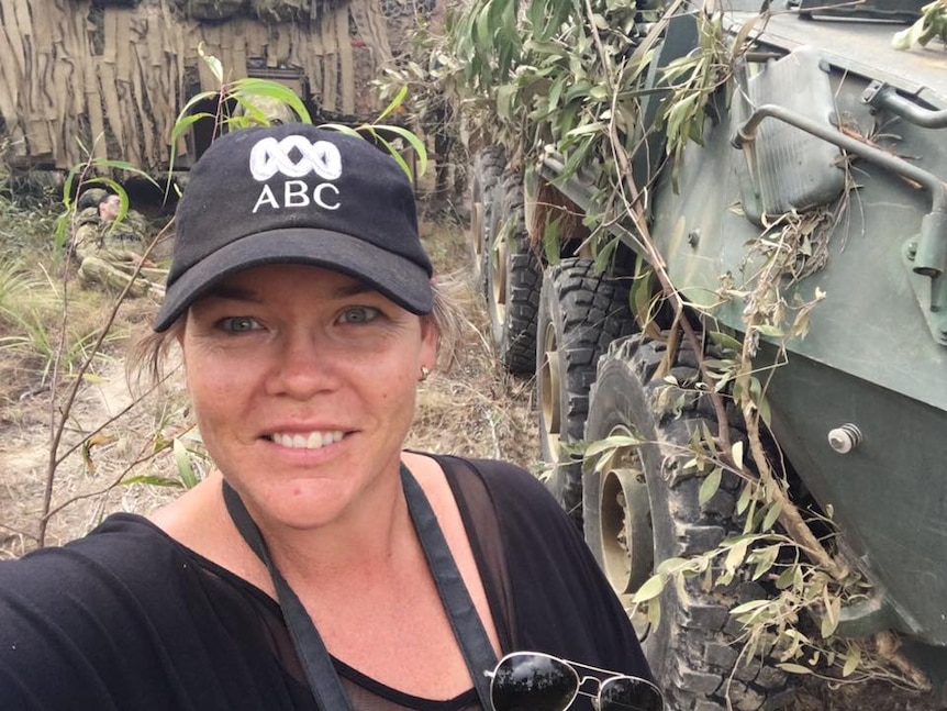 A woman wearing a black ABC cap takes a selfie in bushland with a tank in the background