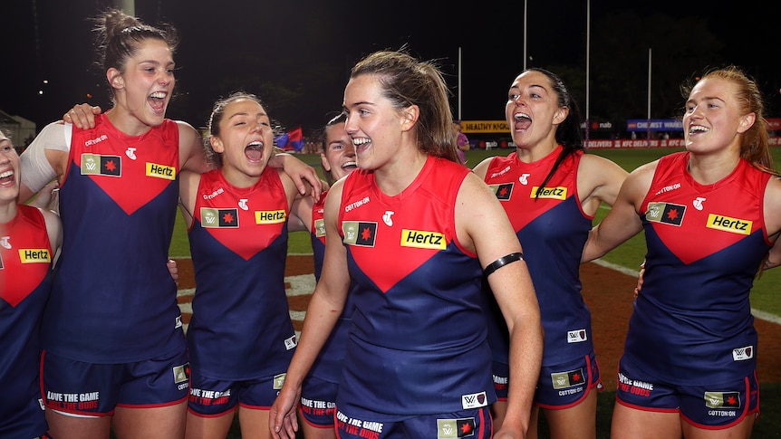 An Irish player in Melbourne's AFLW side stands surrounded by her teammates for the club song after a win.