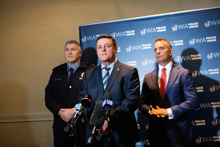 A man in a suit and tie speaks in front of microphones, watched on by two other men