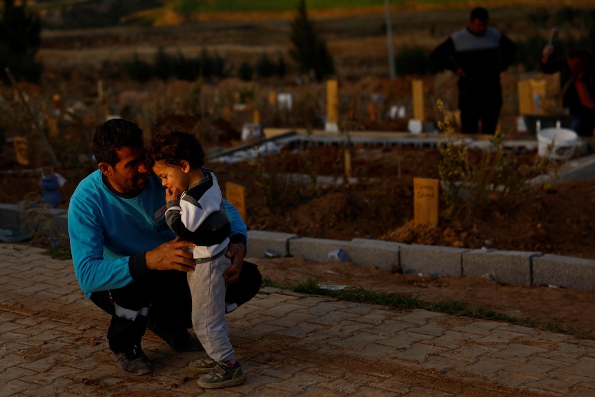 A man crouches down next to his son with graves visable behind them. 