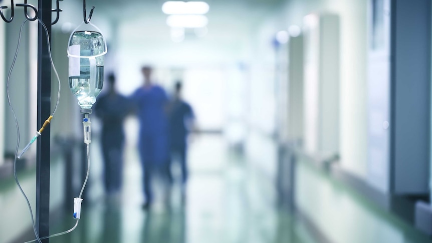 Blurred photo of hospital corridor. Human figures in the backgound.  Saline drip in focus in the foreground