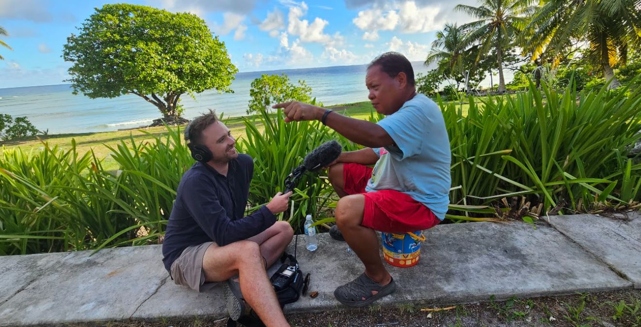 Man, wearing blue shirt and red shorts sits on small bucket surrounded by greenery being interviewed by another man holding mic