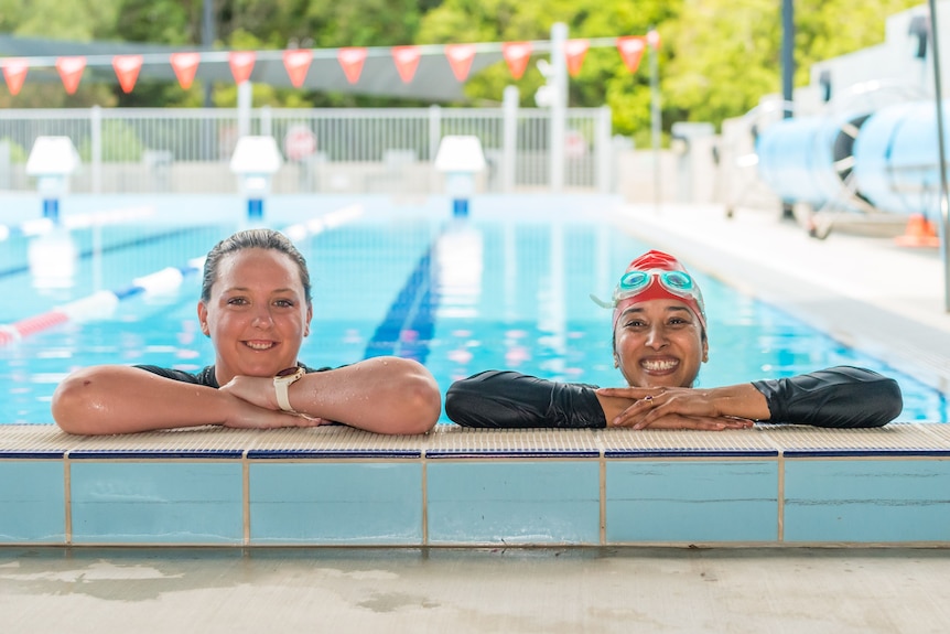 Michelle Campbell Burns and Monika Manot pose for a photo, resting there head on their arms on the edge of a pool.