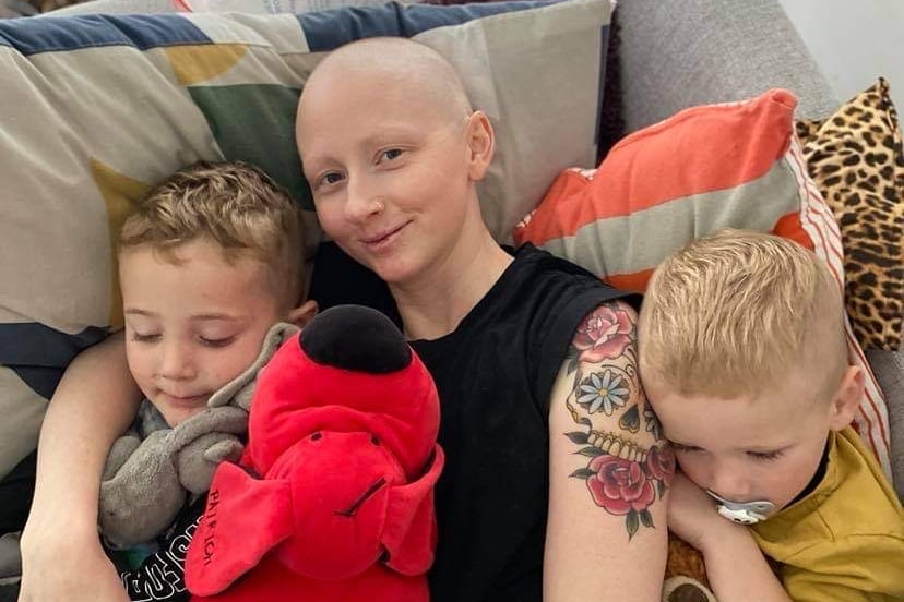 Jodie Leworthy with bald head, lies on a sofa with her two sons