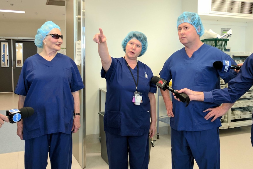 A woman points out features of the new hospital to two other people, all of them in hospital gowns and hats.