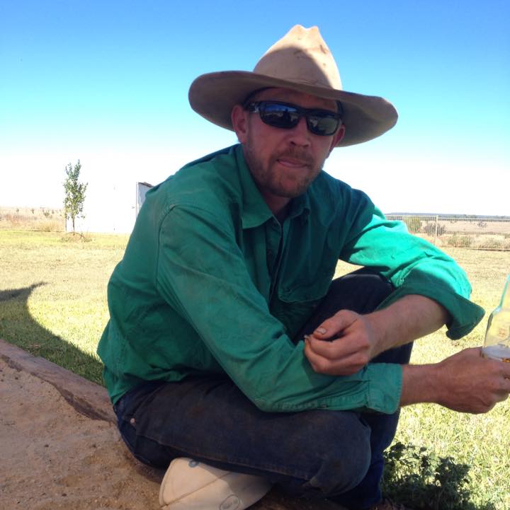 A man wearing a cowboy had and a green shirt sits on the ground holding a glass bottle