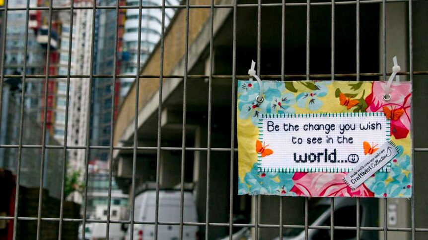 A small stitched sign that reads "Be the change you wish to see in the world" fixed to a construction fence in a city landscape.