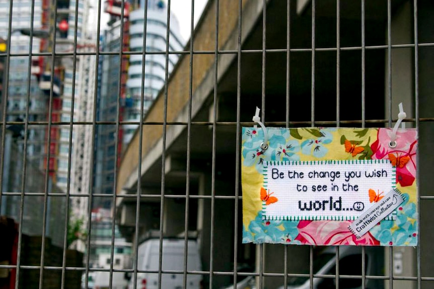 A small stitched sign that reads "Be the change you wish to see in the world" fixed to a construction fence in a city landscape.