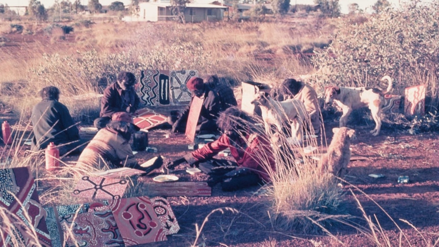 An old photo shows men sitting in red sand painting on wood boards with dogs and spinifex