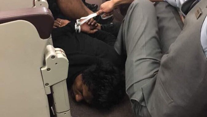 A man is detained on the floor of a plane, held down by a flight attendant.