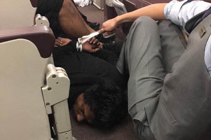 A man is detained on the floor of a plane, held down by a flight attendant.