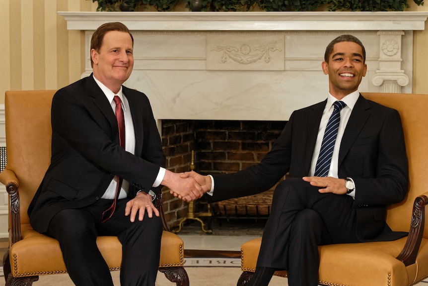Two men shake hands and smile while seated in front of a fireplace