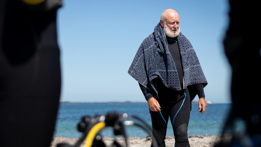 A man with a grey beard wearing a wetsuit stands on the beach with a towel around him.