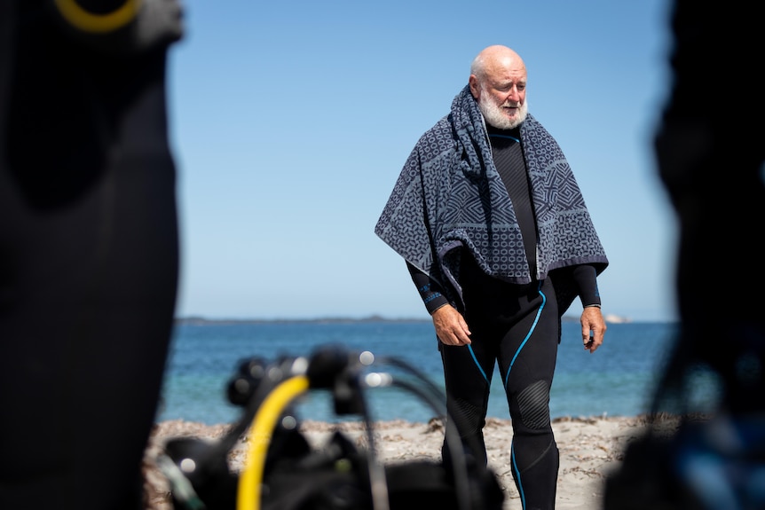 A man with a grey beard wearing a wetsuit stands on the beach with a towel around him.