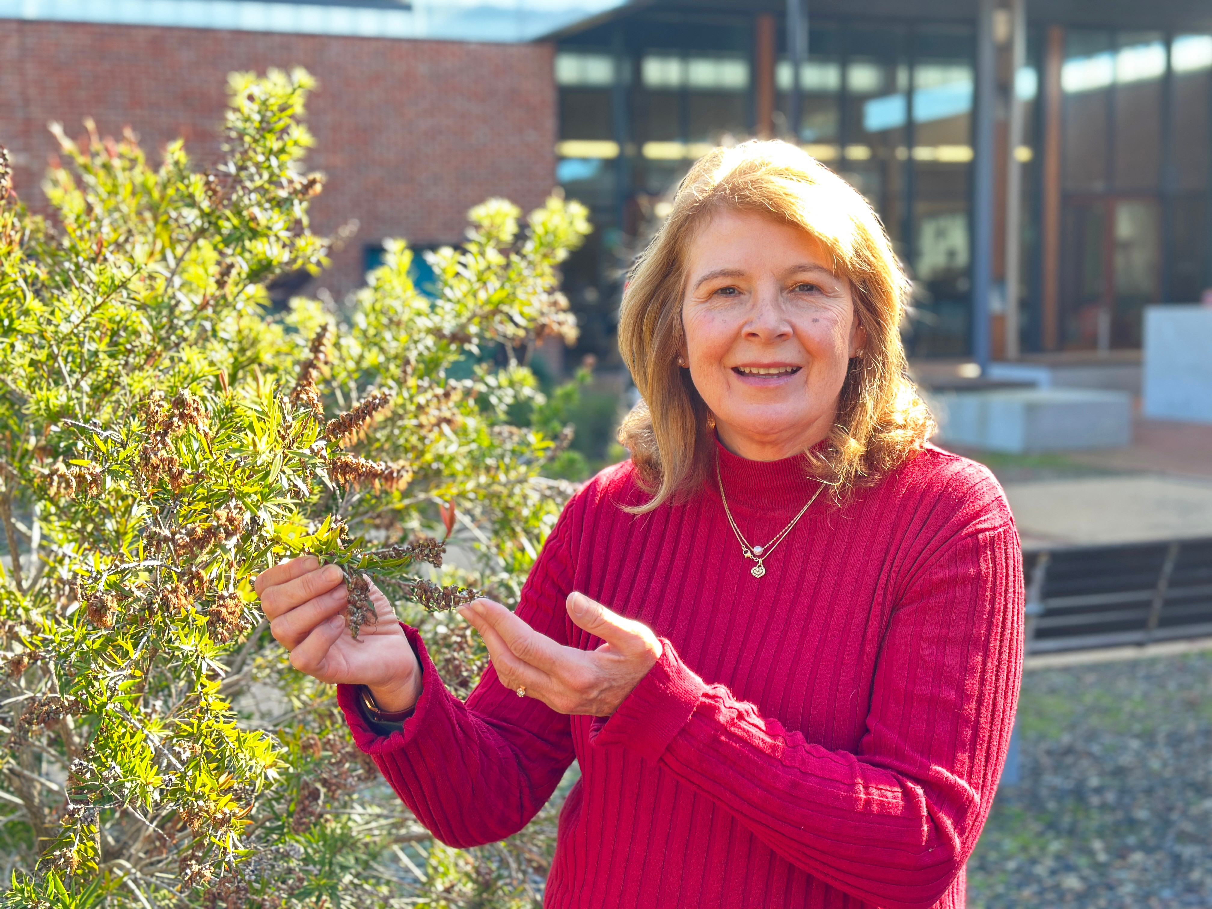 A woman in a red sweater smiling at the camera and pointing out features of a plant.