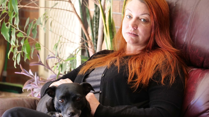 A woman sitting on a dark red leather couch with long bright orange hair and a dog on her lap