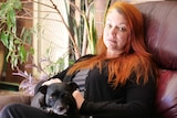A woman sitting on a dark red leather couch with long bright orange hair and a dog on her lap