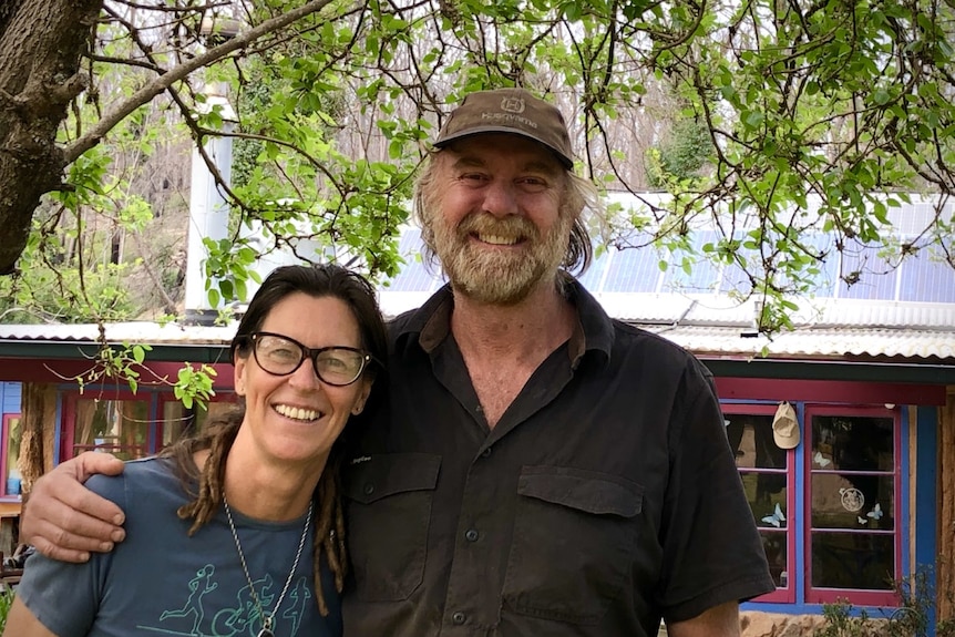 Two people smiling under a big green Mulberry tree in front of a mudbrick house.