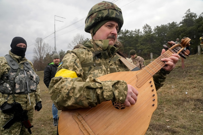 A man in a Ukrainian military uniform plays a kobza string instrument in a field, with another military man standing near him.