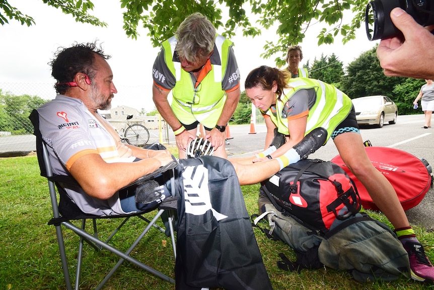 A man in cycling gear sits in a chair while people in high-vis vests massage his legs 