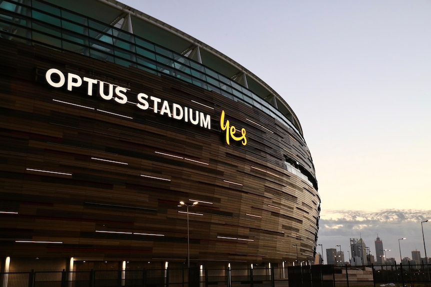 The hulking Perth stadium fills the frame with the Perth city skyline small in the bottom corner at dusk