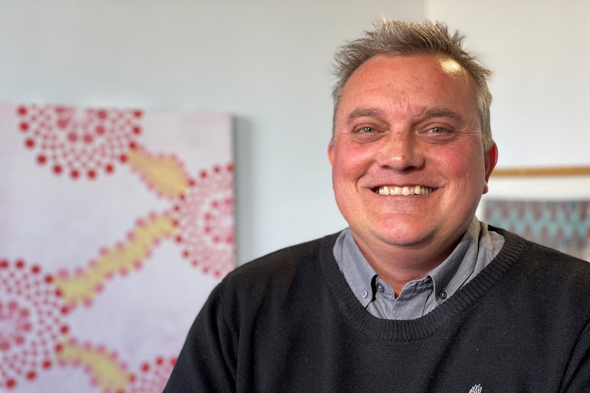 A man with greying hair wearing a grey shirt and dark jumper smiles at the camera