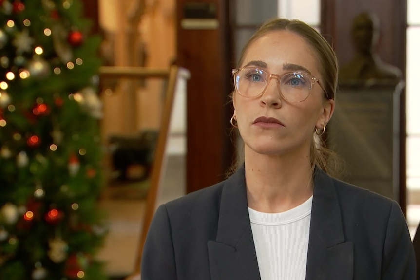 A woman with glasses and hair pulled back stands near a Christmas tree with a serious expression