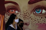 A woman wearing a face mask stands next to a mural of a woman's eyes