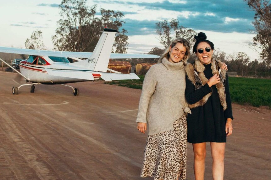 Two women stand smiling in front of a small plane on a runway.