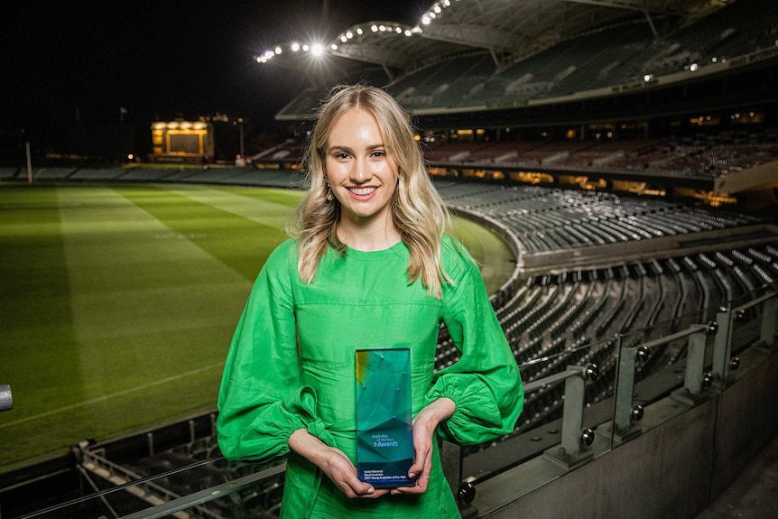 A young woman holds a trophy with a large sports stadium in the background