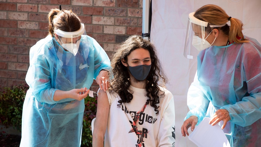 One nurse in full PPE is vaccinating a young man, while another nurse watches over them.