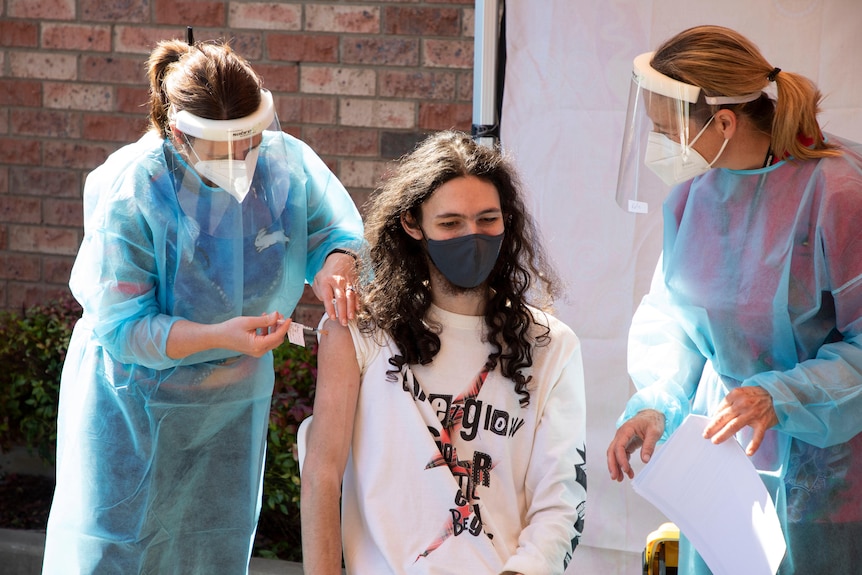 One nurse in full PPE is vaccinating a young man, while another nurse watches over them.