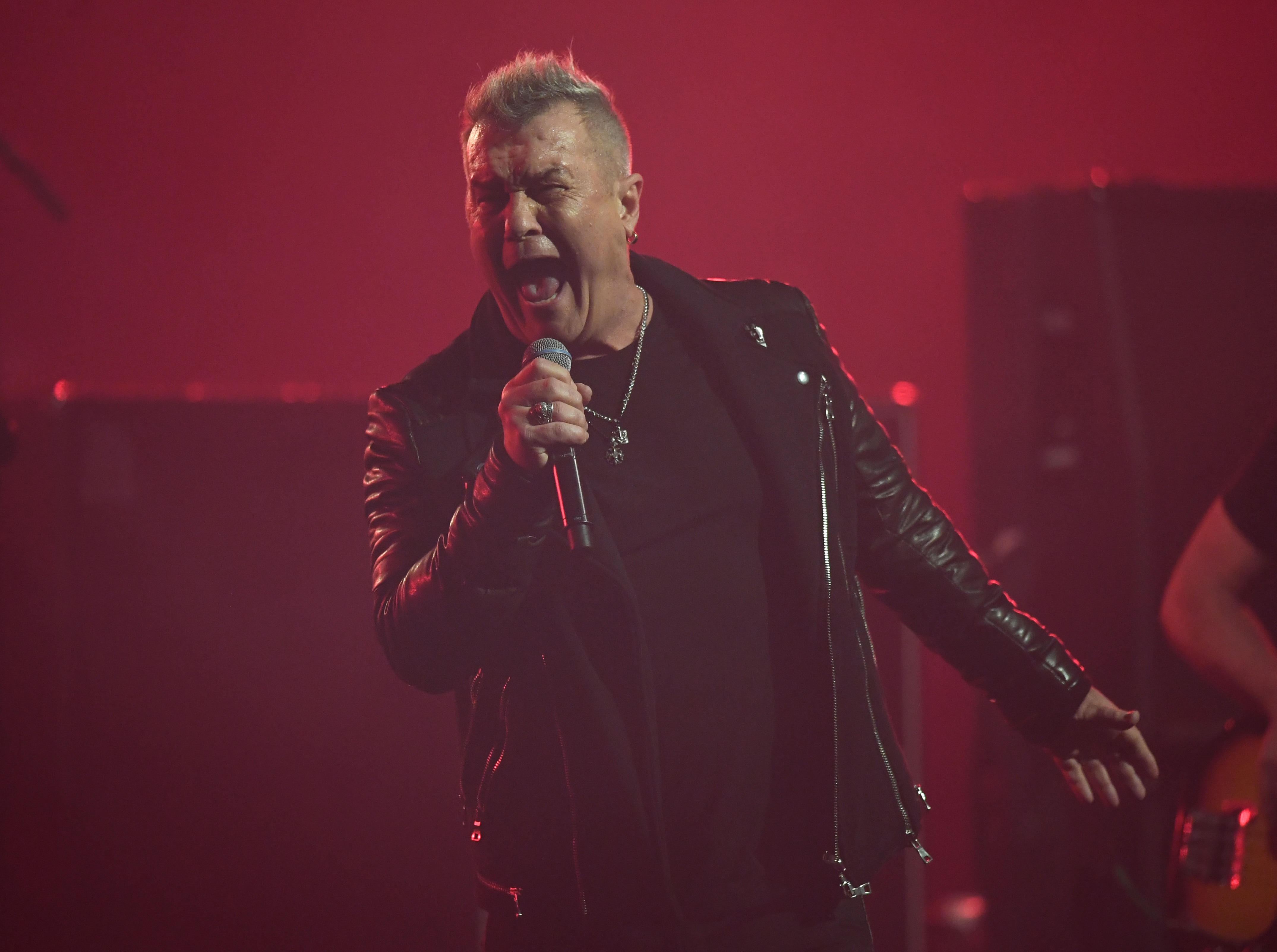 Jimmy Barnes screams into a microphone while wearing a leather jacket onstage.