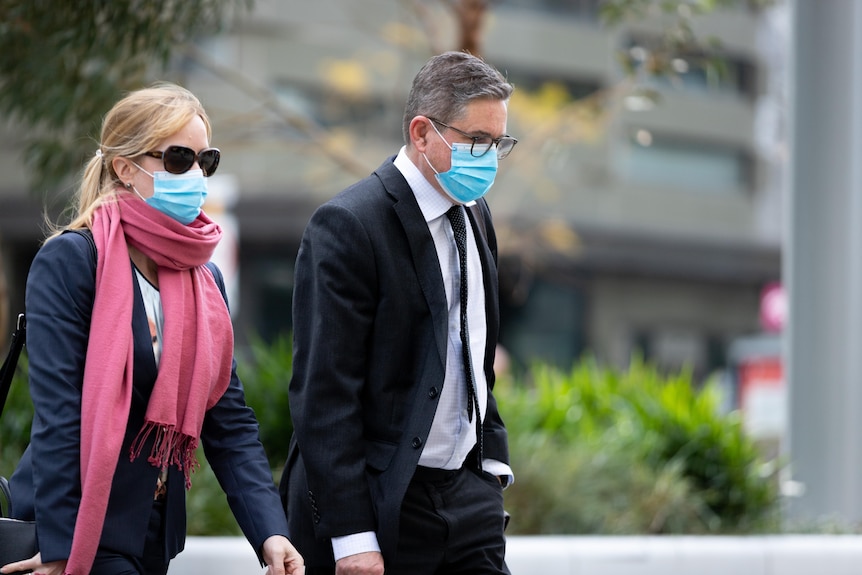 A man in a suit walks next to a woman, both of them wearing face masks