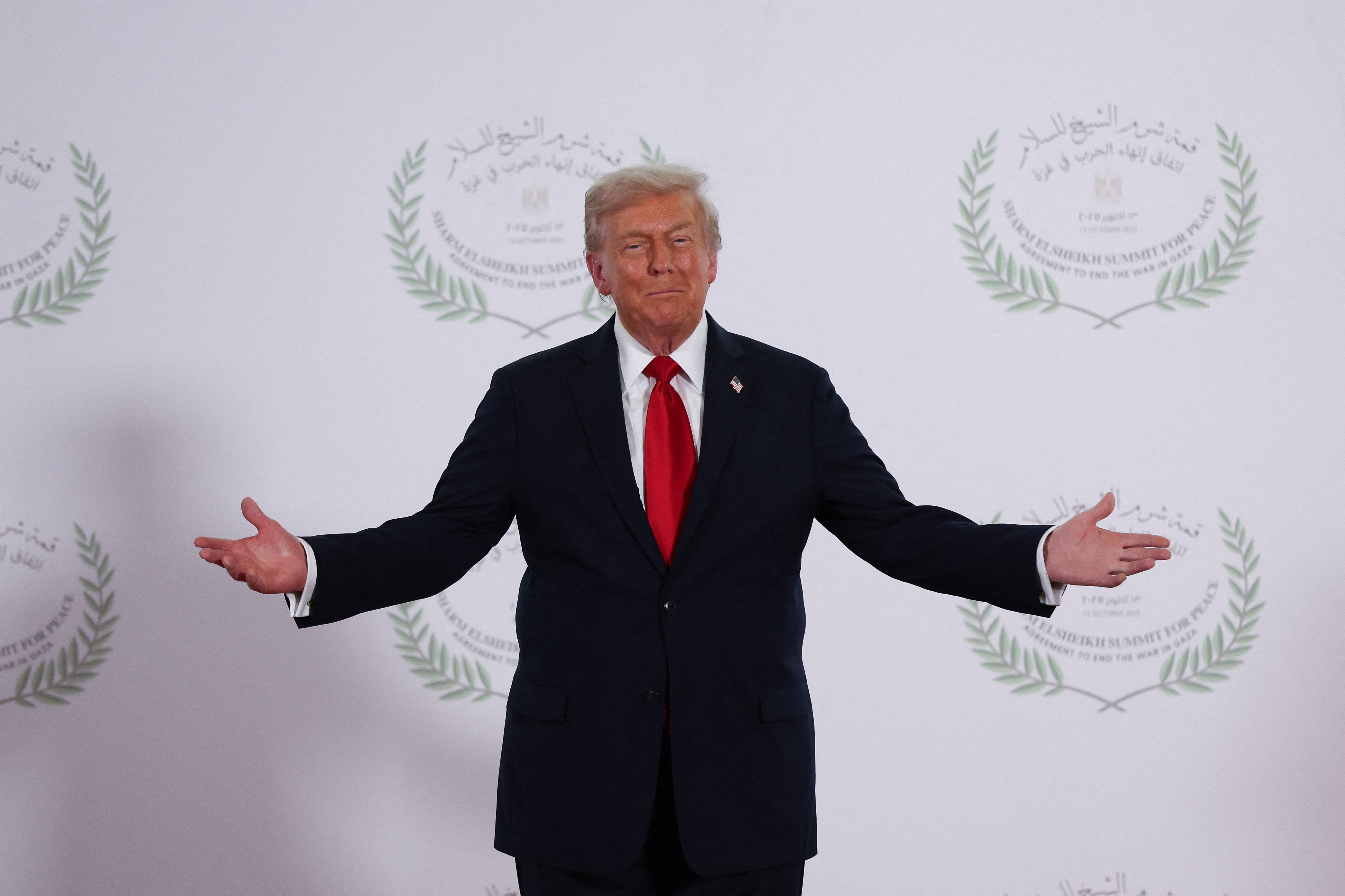 Donald Trump standing in front of a wall with summit logos with his arms wide open