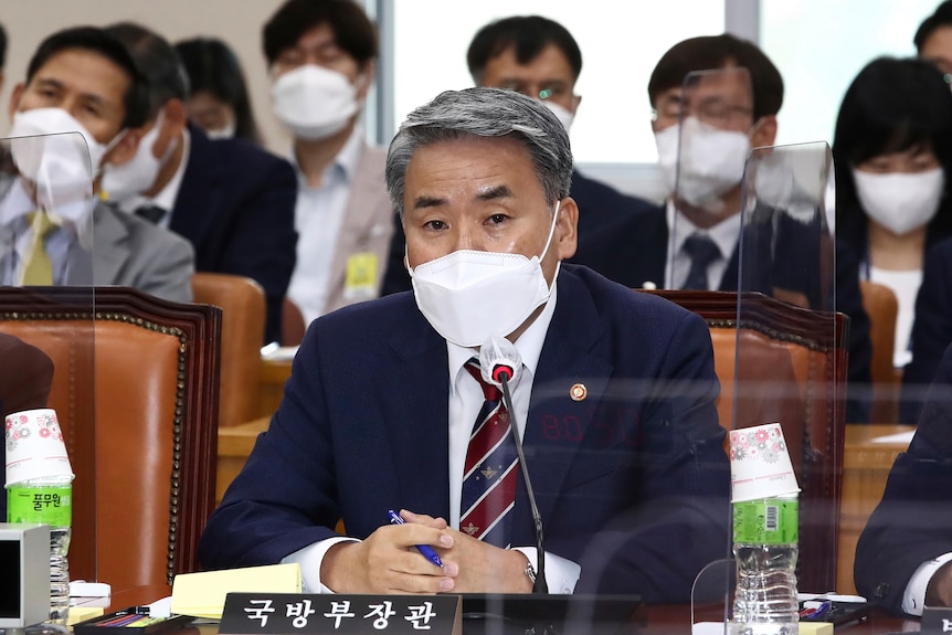 A grey-haired Korean man wearing a navy suit and a surgical mask sits at a table and speaks into a microphone.