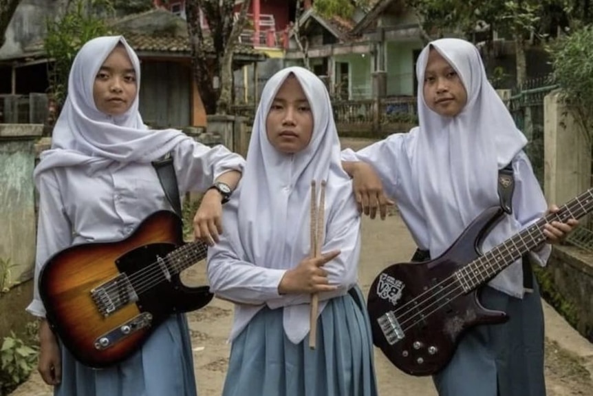 three girls in a white hijab and school uniform hold their instruments, guitar, drumsticks and with rural houses behind them