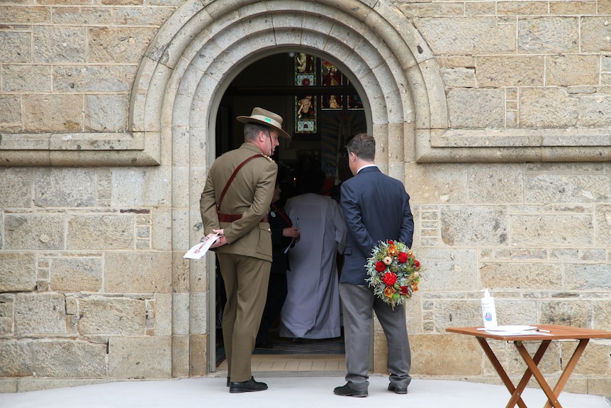 Two men stand looking through the arch stone doorway of a church. One man is dressed in military uniform and one holds flowers.