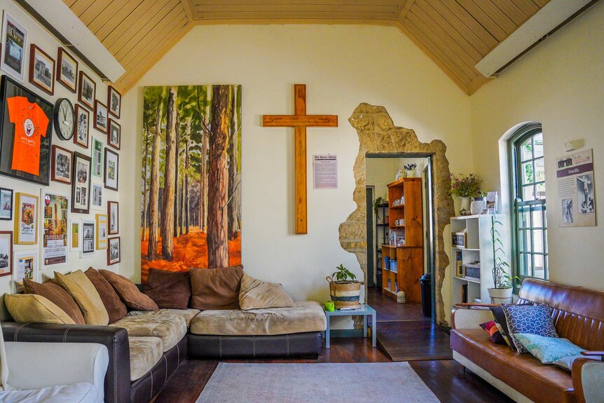 A bi wooden cross is mounted on a wall in a room with comfy lounge chairs and pictures and paintings, and an arched roof