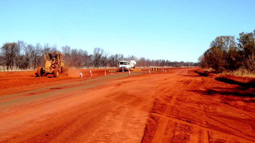 A truck and road work machinery to the left and in the distance. Red road with markers and bushland surrounds.