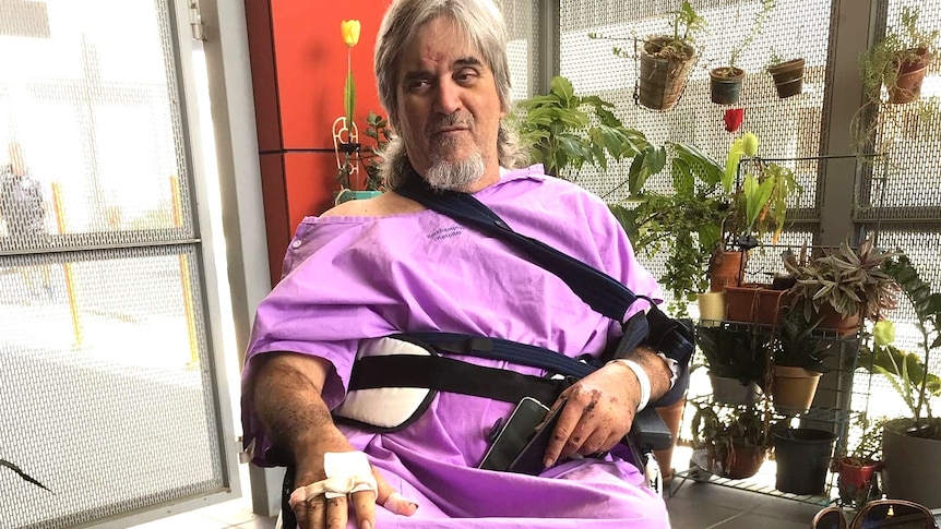 A man wearing a purple hospital gown with his arm in a sling and in a wheelchair.