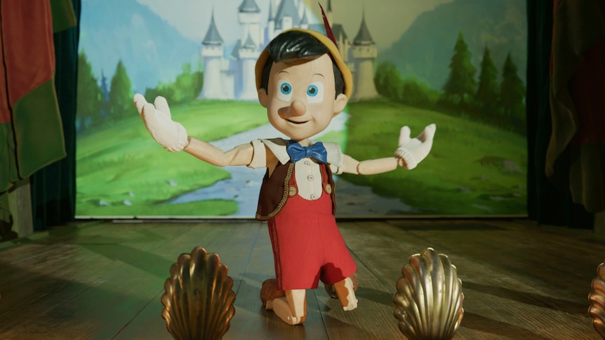 Humanoid toy on a stage performing, smiling, arms apart, wearing red overalls with brown vest and white top and gloves.