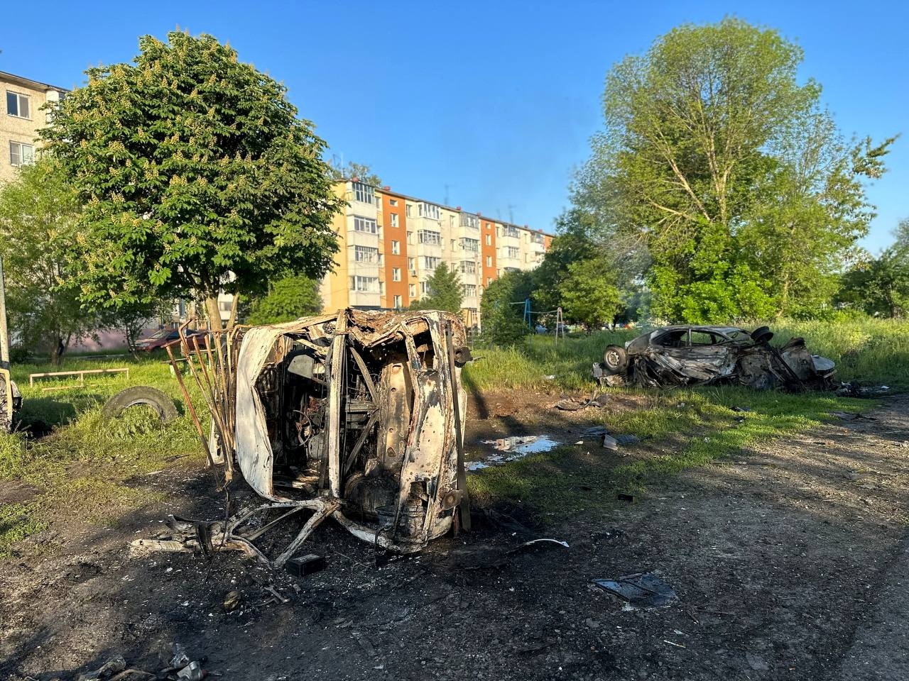 Destroyed vehicles sit next to a dirt road with residential buildings in the background.
