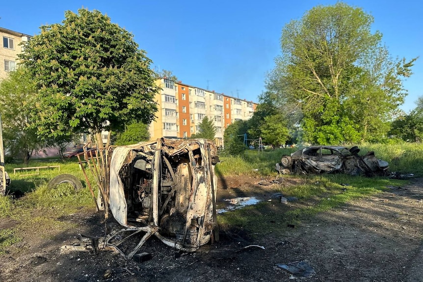 Destroyed vehicles sit next to a dirt road with residential buildings in the background.